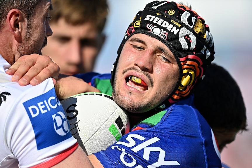 A Warriors NRL player holds the ball against his chest as he is tackled by a Dragons opponent.