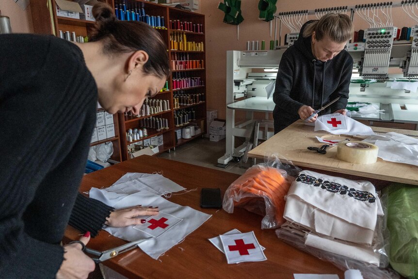 Two women work at tables as they cut out red crosses on material.