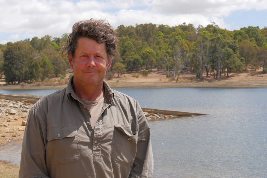 A man in a khaki shirt looks at the camera with a lake in the background.