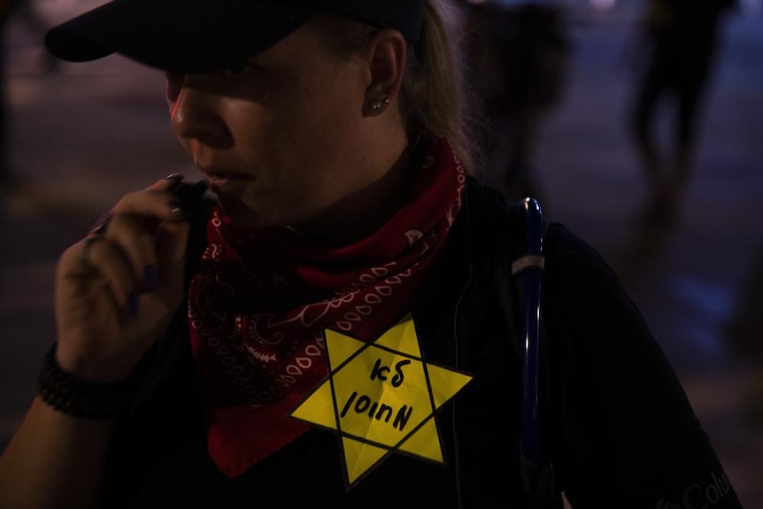 A woman wears a yellow Star of David on her black t shirt in a dimly lit street at night 