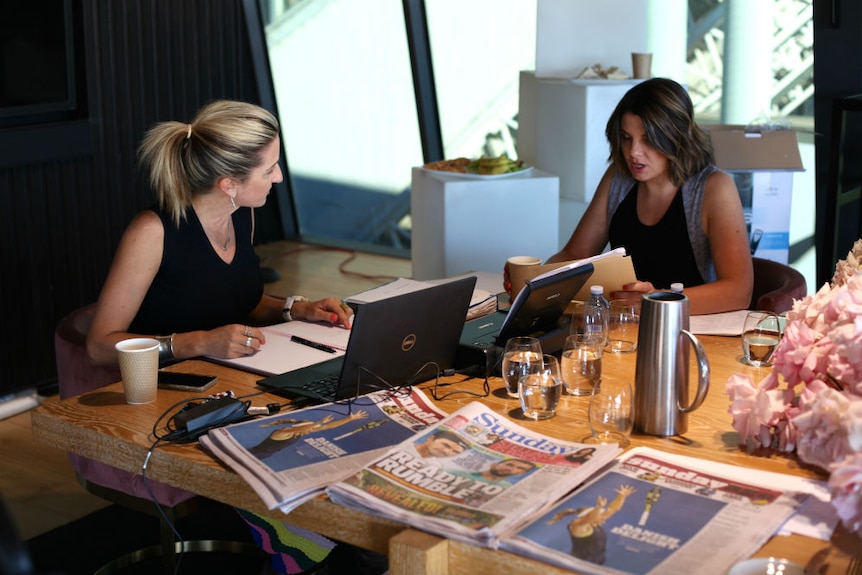 Kate Tozer and Kelli Underwood sitting in front of lap tops with newspapers on table.