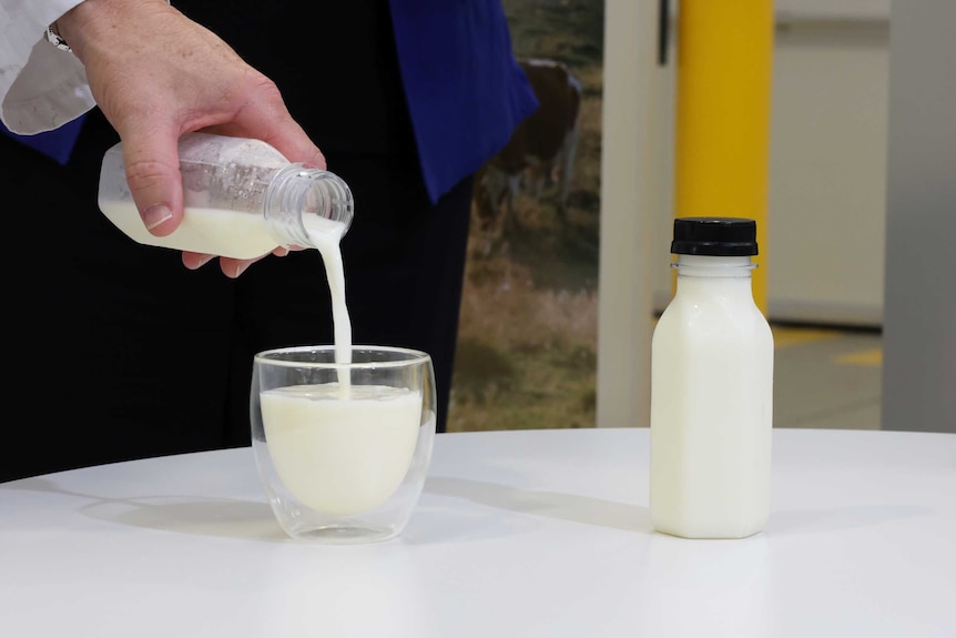 Hand in a lab coat sleeve pours milk from an unlabelled bottle into a small glass on a white table.