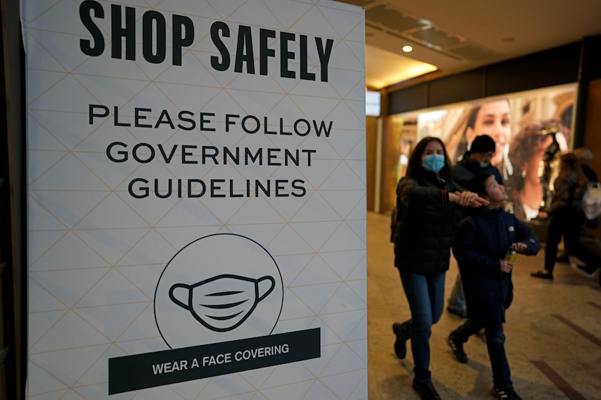 A family of three with a young boy walking past a sign with the words 'Shop safely' and COVID-19 guidelines