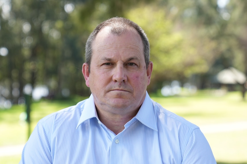 A balding man in an open-neck blue business shirt looks serious as he poses for a photo in a park.