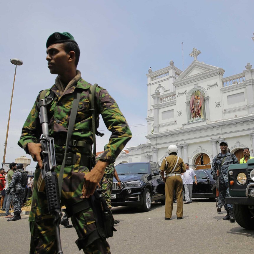 From a low angle, you look up at a soldier in camouflage with a large automatic gun in front of a white neo-classical church.