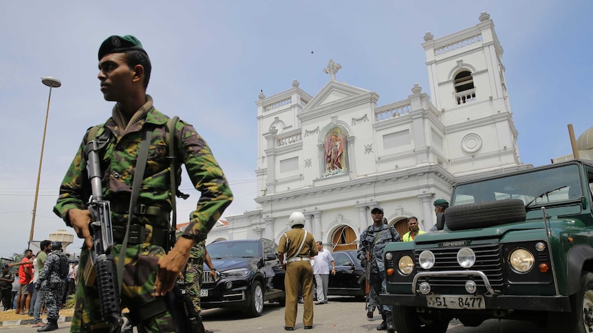 From a low angle, you look up at a soldier in camouflage with a large automatic gun in front of a white neo-classical church.