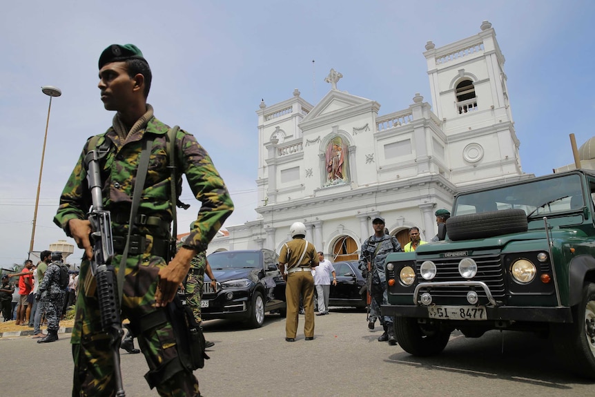 From a low angle, you look up at a soldier in camouflage with a large automatic gun in front of a white neo-classical church.