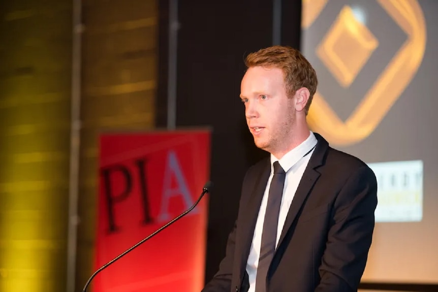 A man in a black and white suit and tie, standing at a lecturn, speaking into a microphone.