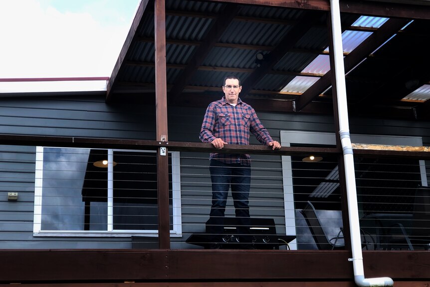 A man is standing on a deck in-front of a dark grey house looking into the camera