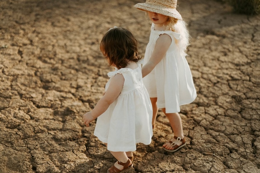 Two toddler-aged girls in white dresses hold hands while walking on the cracked earth of a dry and barren paddock.
