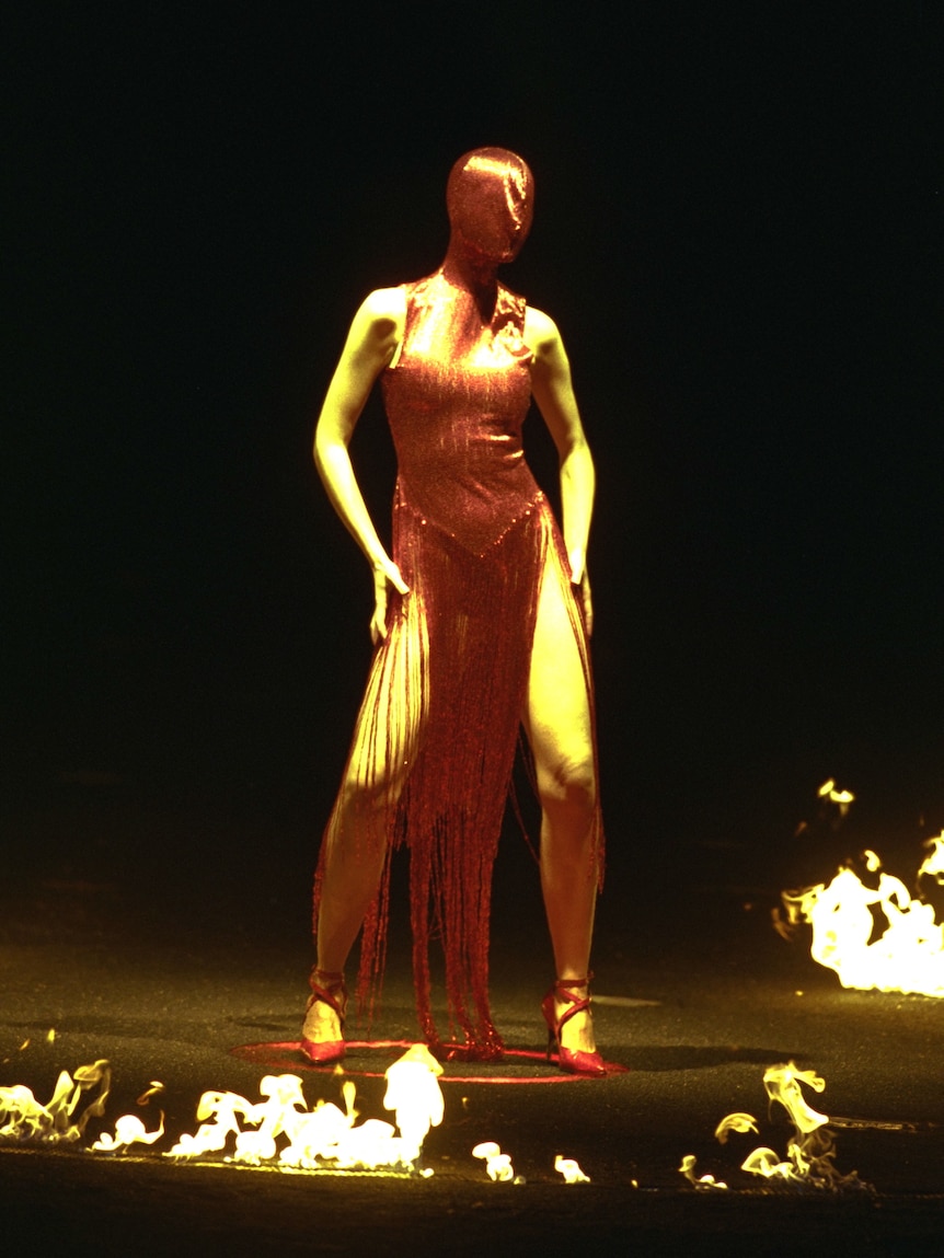 Model wearing red shiny dress covering entire head stands on dark stage with a ring of fire around her.