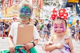 Two young girl with painted faces hold showbags in front of large ferris wheel