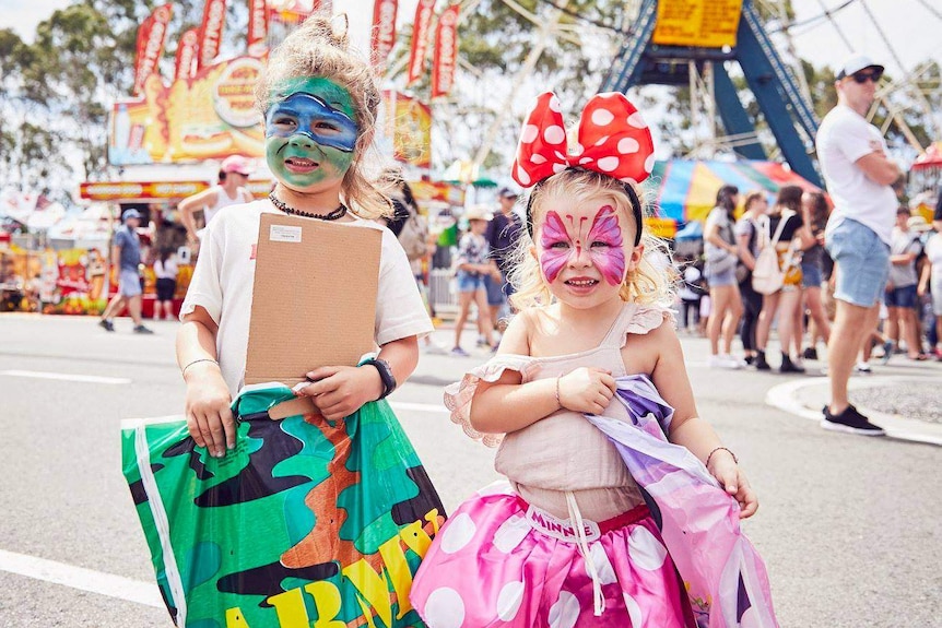 Two young girl with painted faces hold showbags in front of large ferris wheel