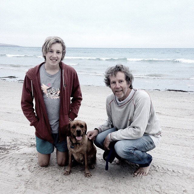 A teenaged boy with a small brown dog and man crouch down on the beach in front of water