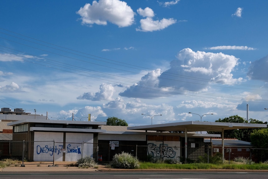 An empty building with a steel fence around it and graffiti on the outside, with blue sky and clouds rolling into the distance. 