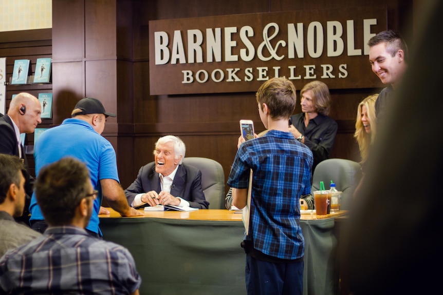 An elderly man sits behind a desk at a book signing, greeting fans.