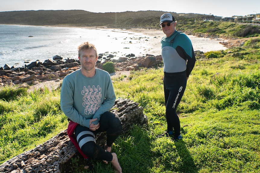 Two men wearing wetsuits on grass near a beach.