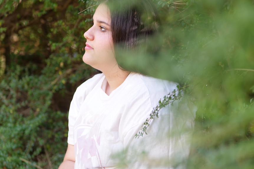 A teenage girl wearing a light pink t-shirt sits among trees looking off to the right. 