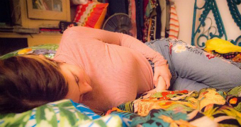 A woman holds her stomach as she lies on multicoloured bed spread with pink top and grey jeans on.