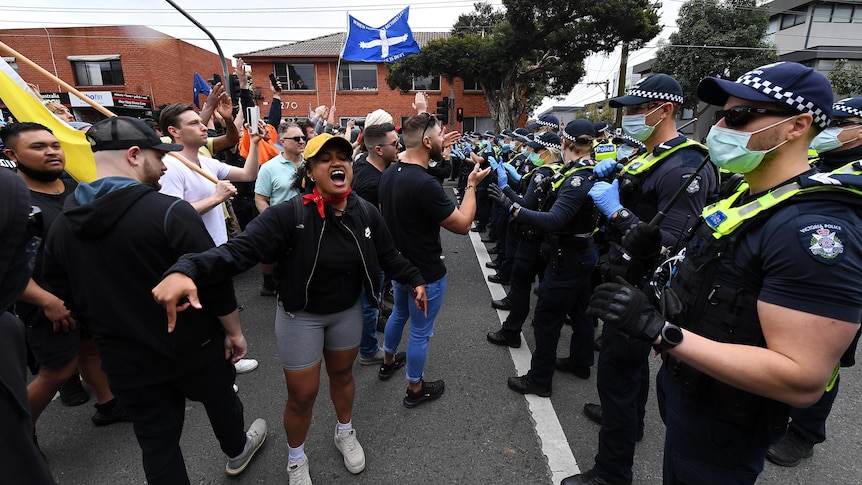 Maskless protesters stand in a line opposing a row of police, with some protesters yelling and waving flags.