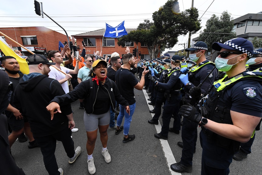 Des manifestants sans masque font la queue face à une rangée de policiers, certains criant et agitant des drapeaux.