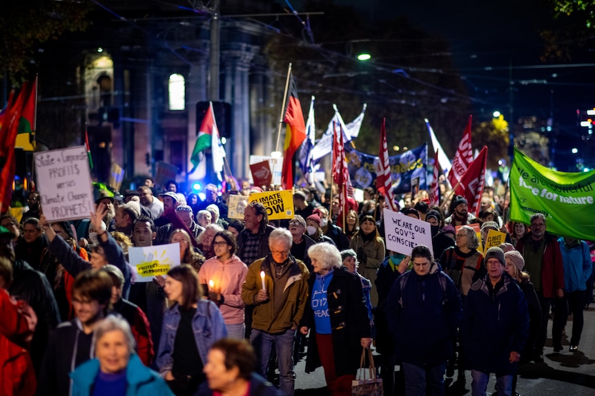 Protesters hold signs, flags and candles while marching on a street at night.