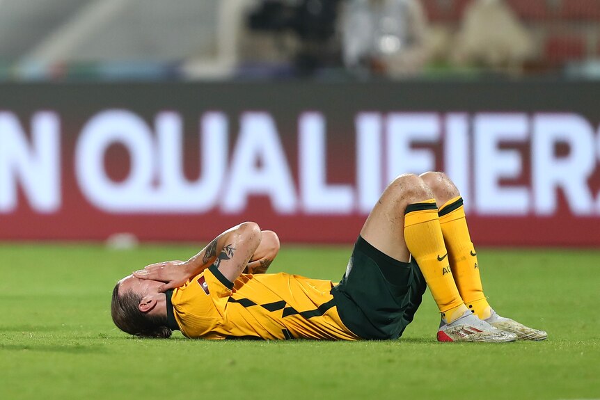 Soccer player wearing yellow and green uniform lays on his back in the grass with his face in his hands