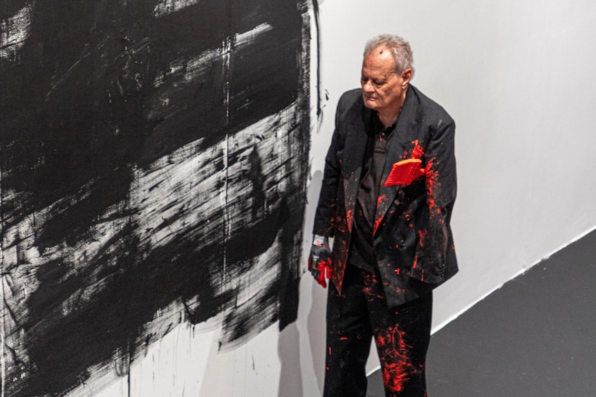 A man in a black suit with his eyes closed is splattered in red paint next to a wall splattered in black.