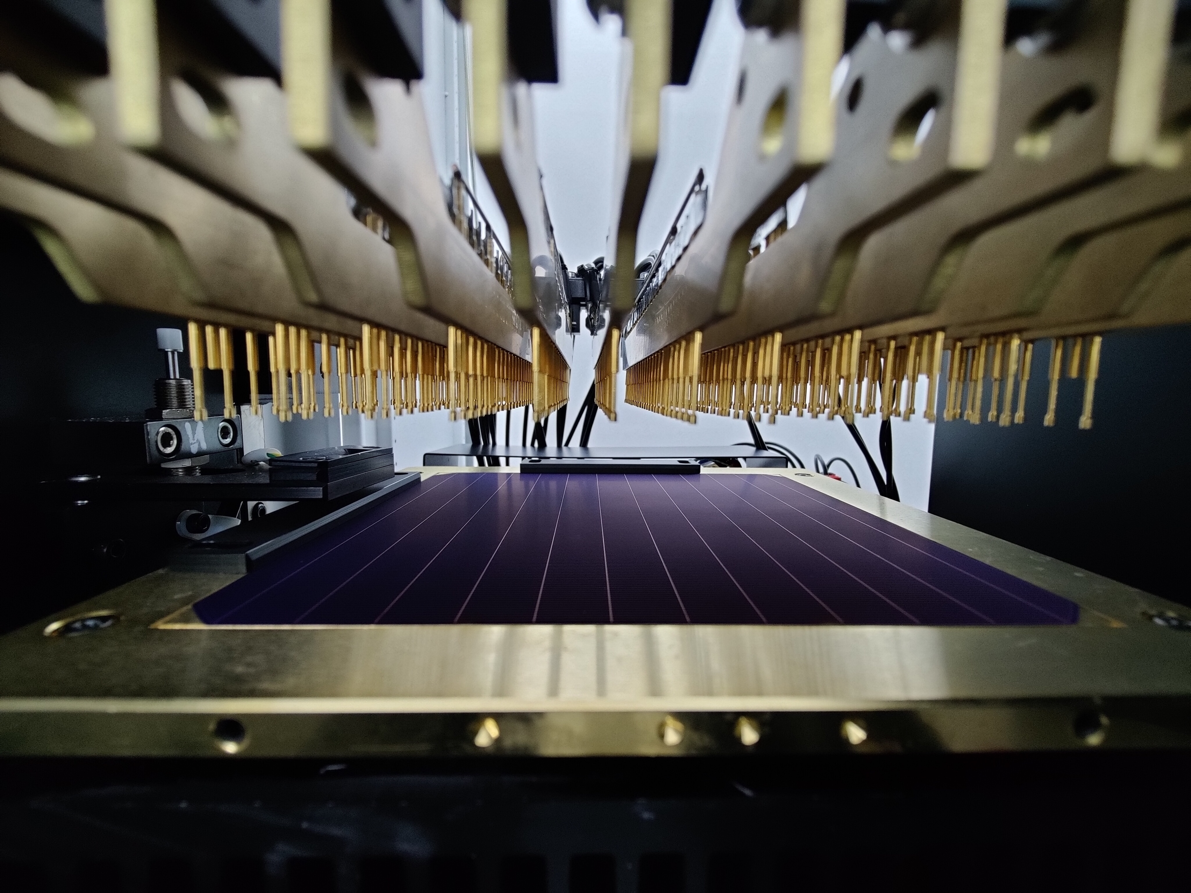 A blue solar cell sits on a platform inside a laboratory underneath a number of gold metal pegs.