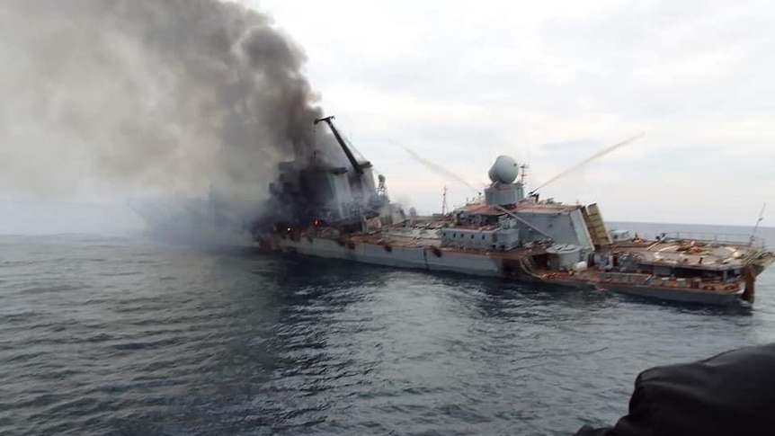 Smoke billows from a tilted warship on a grey sea, as viewed from a ship nearby