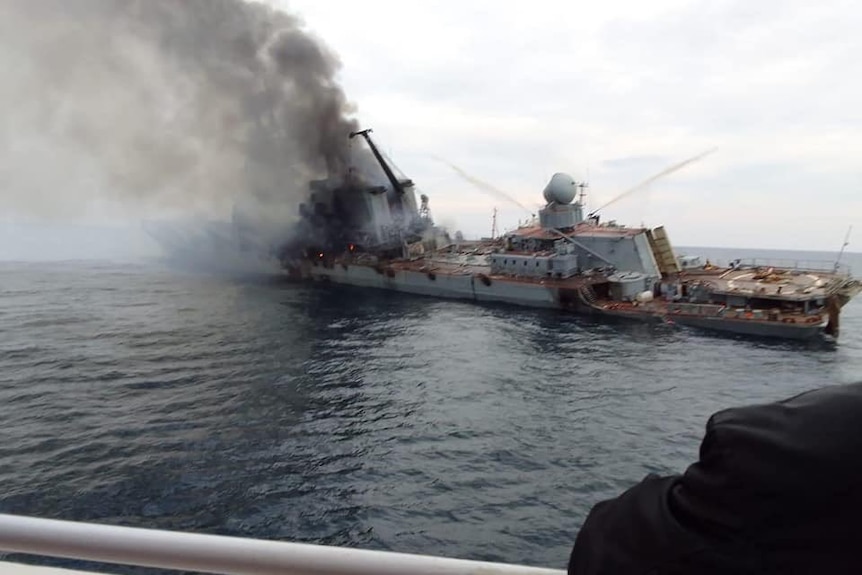 Smoke billows from a tilted warship on a grey sea, as viewed from a ship nearby