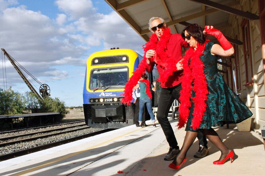 A man and a woman in red feather boas dance on a train platform.
