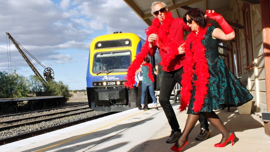 A man and a woman in red feather boas dance on a train platform.