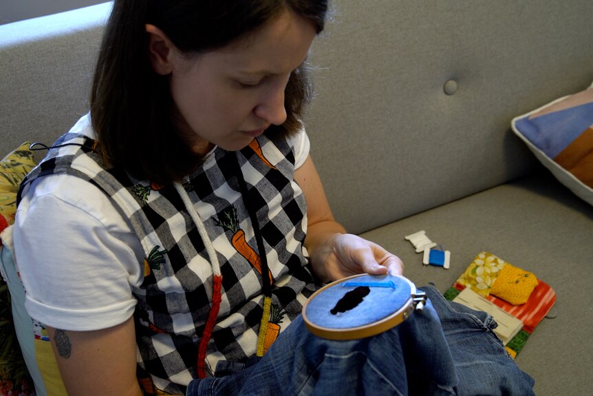 Renae sitting on a couch repairing a hole in a pair of blue jeans using blue thread