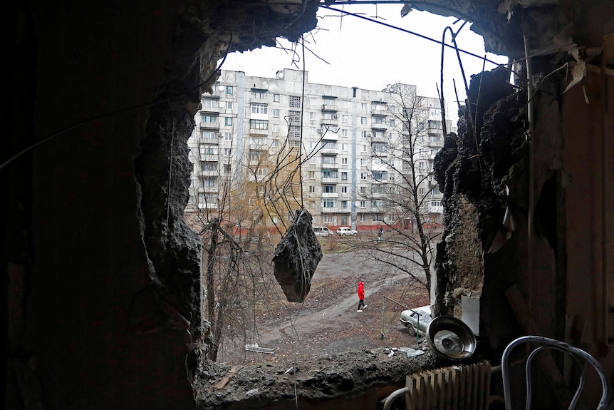 Through a hole in a wall of a building, a person is seen walking down a path outside.