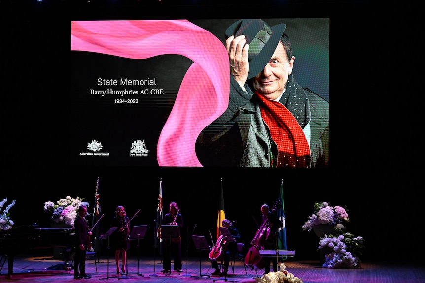 An image of late entertainer Barry Humphries beams from a large screen at his memorial service