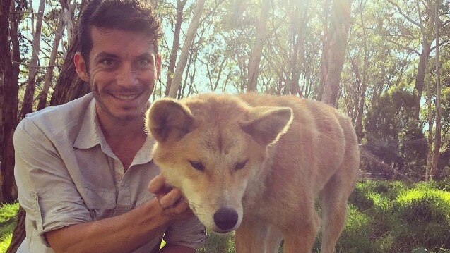 A man with black hair smiles at the camera while patting a dingo