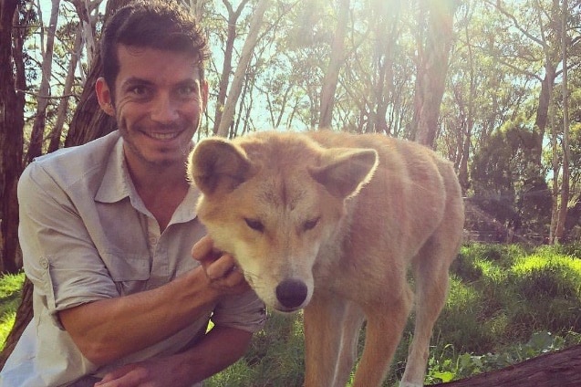 A man with black hair smiles at the camera while patting a dingo