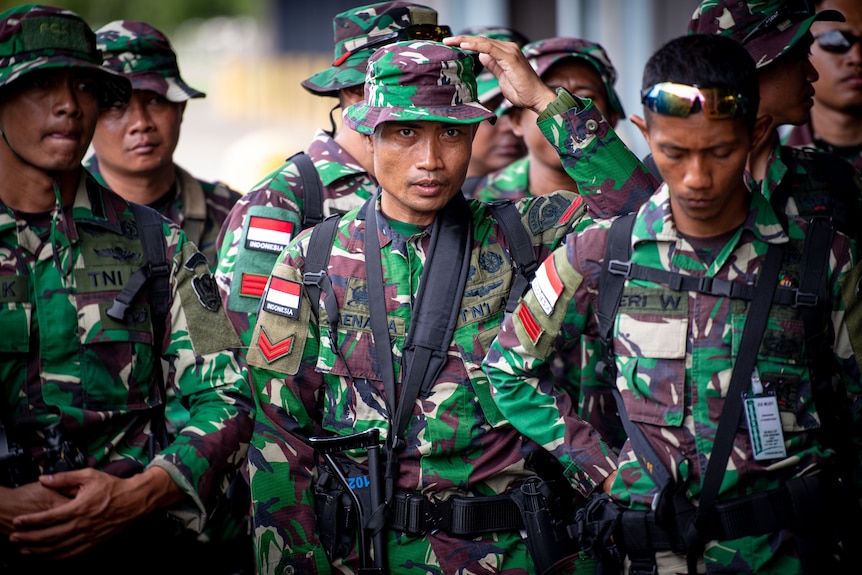 Indonesian soldiers, one wearing a camouflage bucket hat, standing in a group.