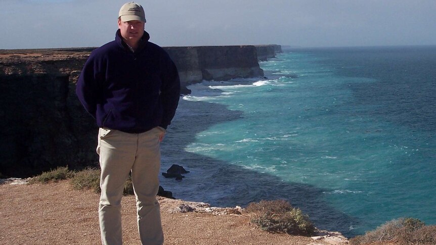 A man in a cap stand in front of some large sea cliffs