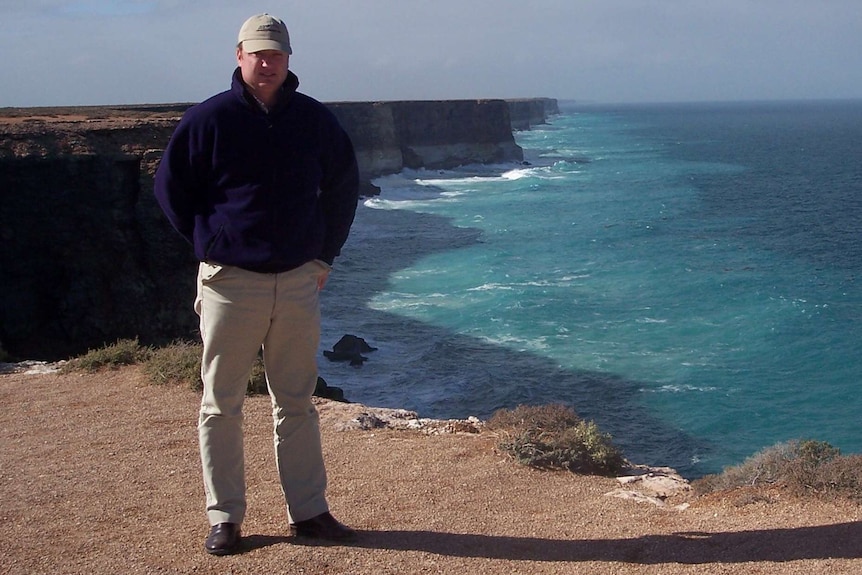 A man in a cap stand in front of some large sea cliffs