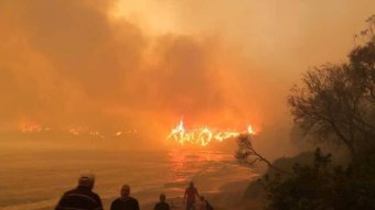 People walk onto a darkened beach as flames can be seen burning on a beach in the distance.