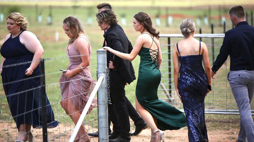 A group of year 12's in colourful dresses and suits walk through a farm gate on a paddock chatting.