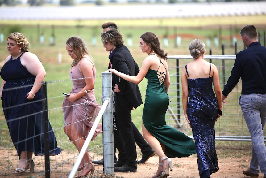 A group of year 12's in colourful dresses and suits walk through a farm gate on a paddock chatting.