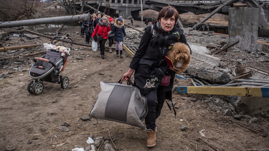 A woman holding a small dog and a bag walks through rubble near an empty pram, as a group of people walk behind her.