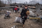 A woman holding a small dog and a bag walks through rubble near an empty pram, as a group of people walk behind her.