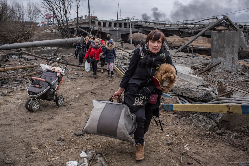 A woman holding a small dog and a bag walks through rubble near an empty pram, as a group of people walk behind her.