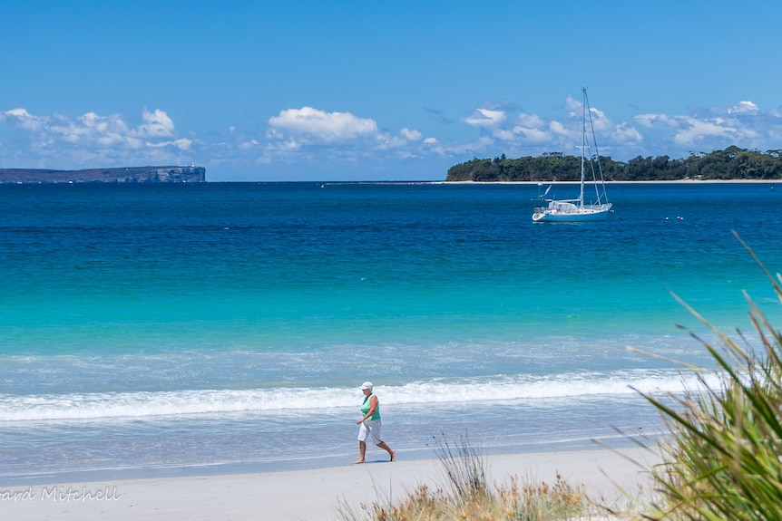 A woman enjoys a walk along Vincentia Beach on the NSW south coast, as  yacht is moored off the beach.