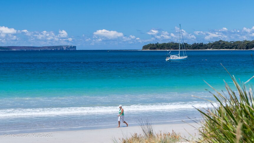 A woman enjoys a walk along Vincentia Beach on the NSW south coast, as  yacht is moored off the beach.