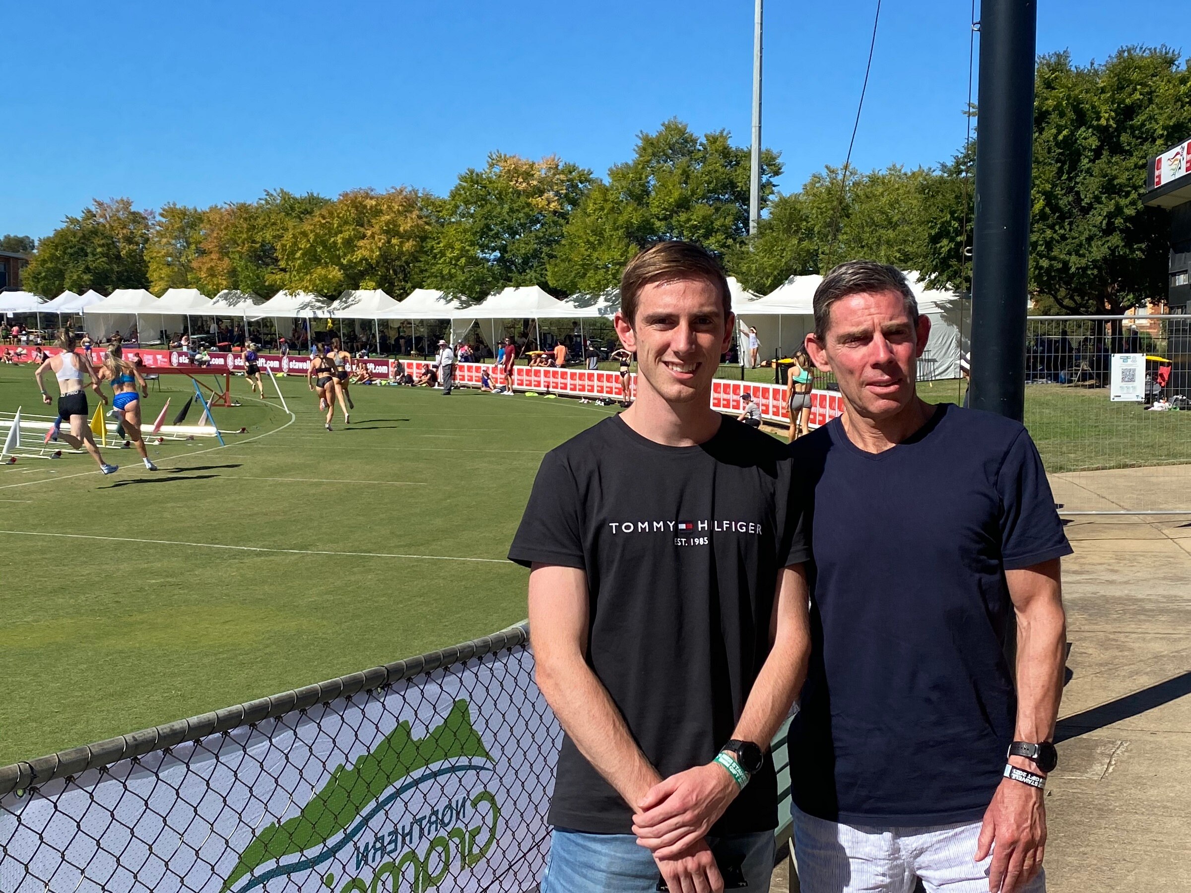 A young man and an older man stand beside an athletics track where women are running.